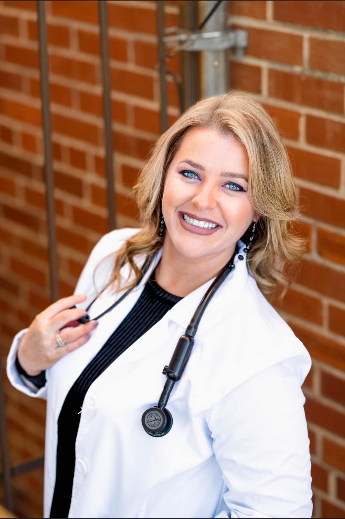 A woman in a lab coat with a stethoscope around her neck is smiling in front of a brick wall.