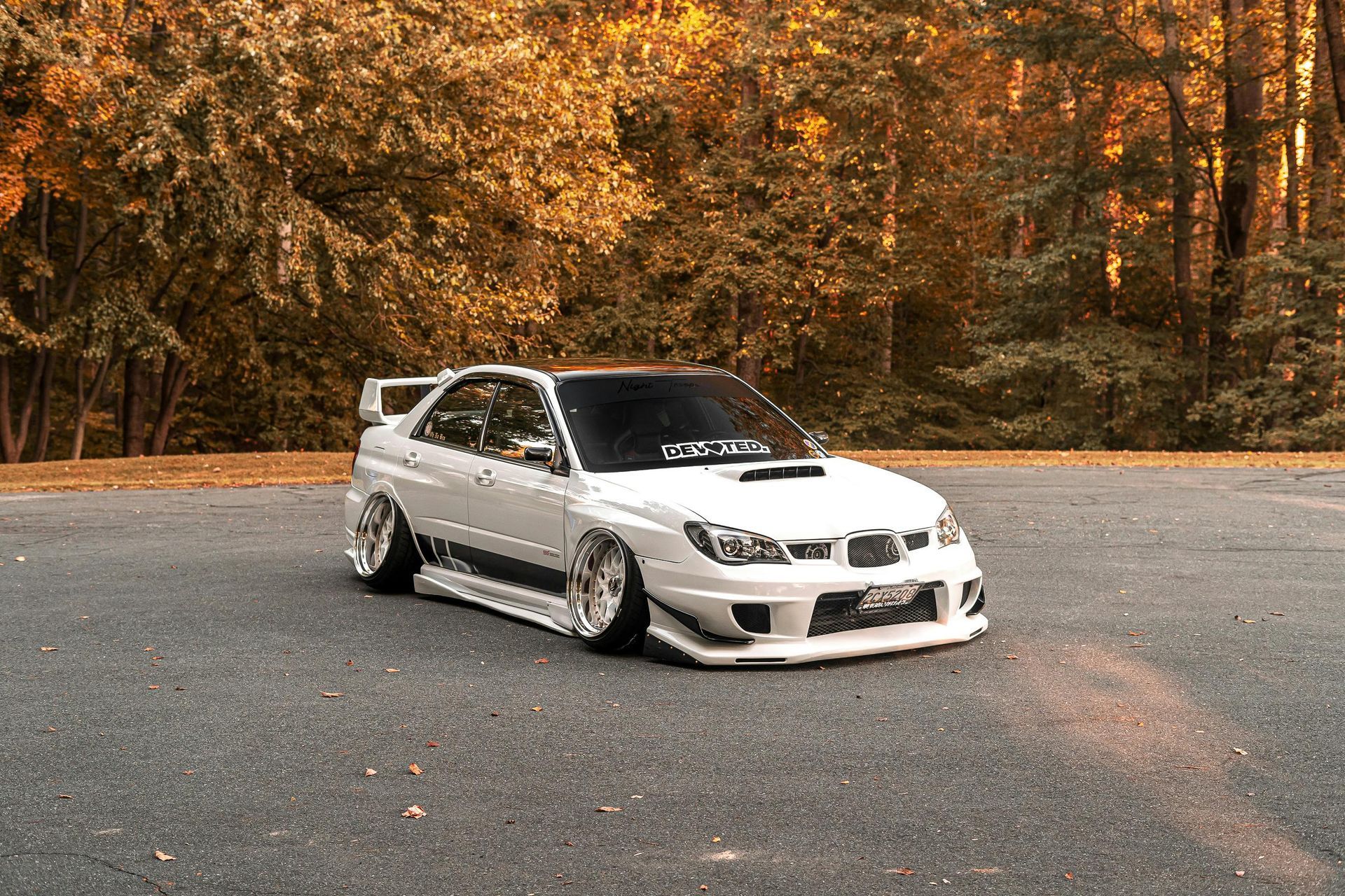 White Subaru Impreza with modified body kit, lowered suspension, parked on asphalt with fall trees in background.