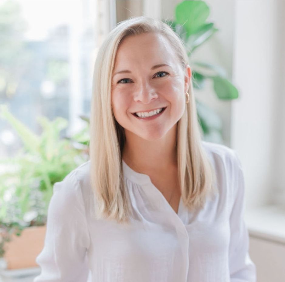A woman in a white shirt is smiling in front of a window.