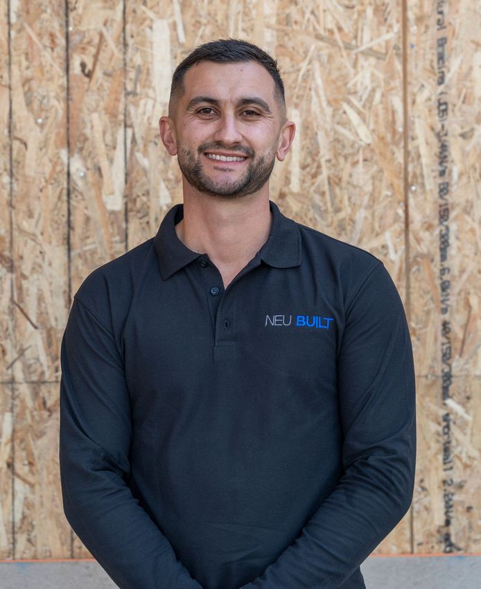 Spero Koulianos of Neu Built wearing a black long-sleeved polo shirt, standing in front of a wood wall.