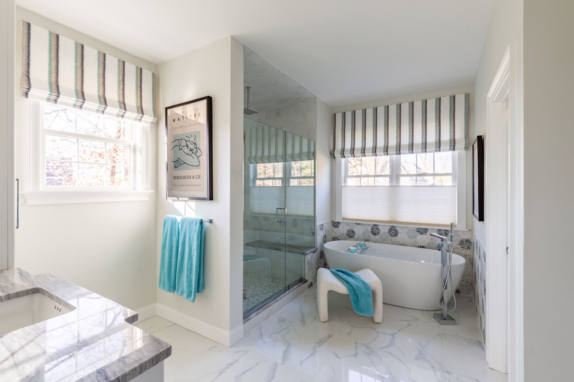 Bathroom with a white tub, glass shower, blue towels, and striped shades; bright and airy.