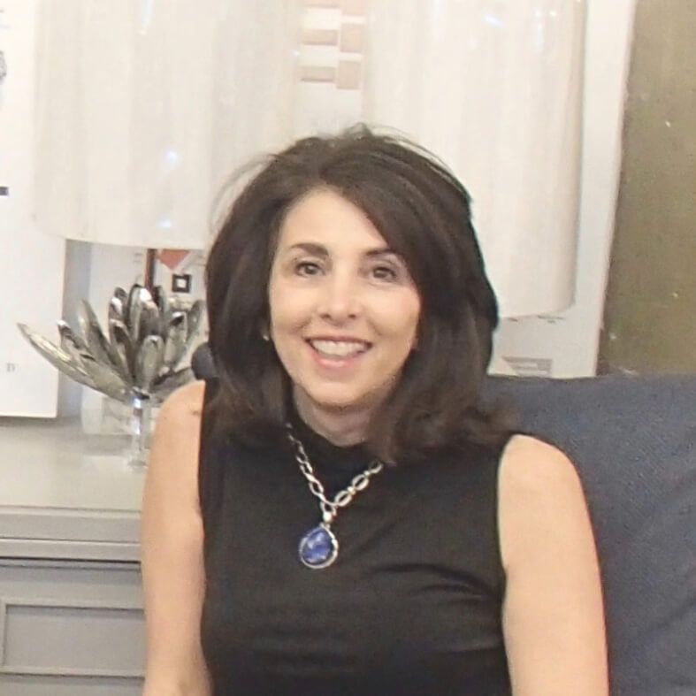 Woman with brown hair, smiling, wearing black top and blue pendant, seated indoors.