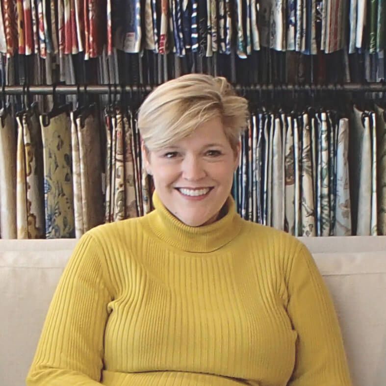 Woman with short blonde hair smiles, wearing a yellow turtleneck in front of fabric samples.