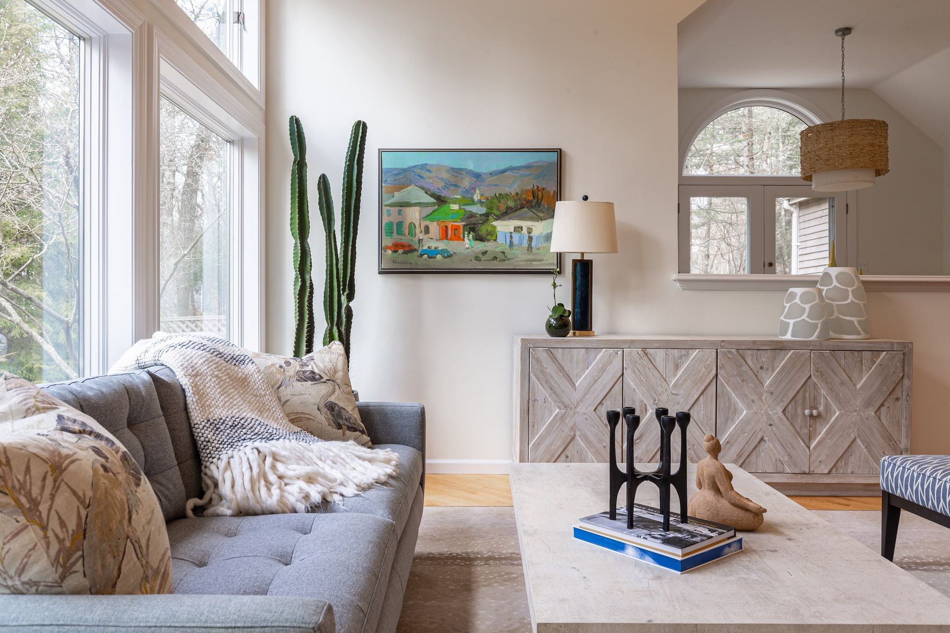 Living room with gray sofa, cactus, art, and a light wood sideboard, with large windows.
