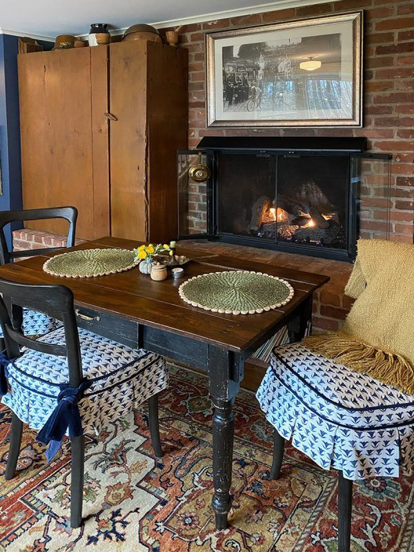 Dining table with two chairs, placemats, fireplace, and cabinet in cozy room with brick and patterned rug.
