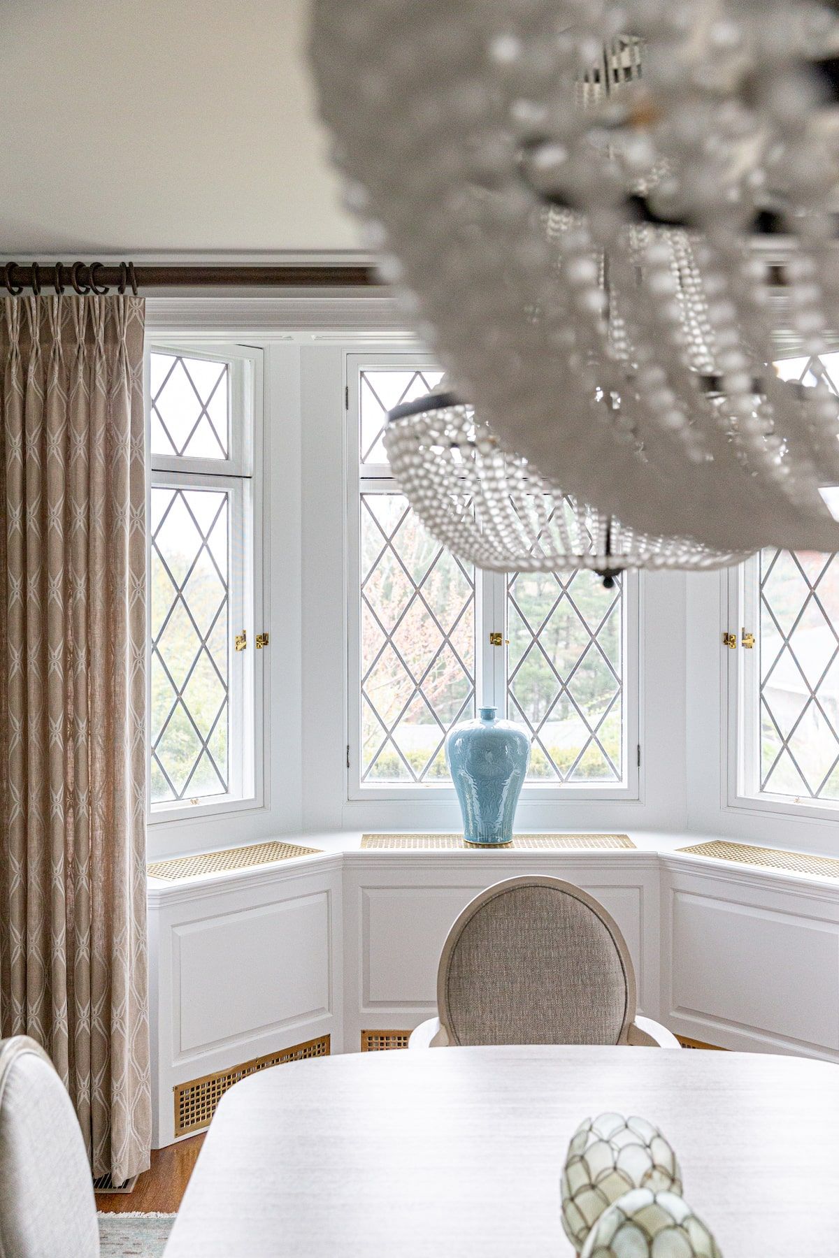 Dining room with bay window, blue vase, white table, beaded chandelier, and neutral-toned curtains.