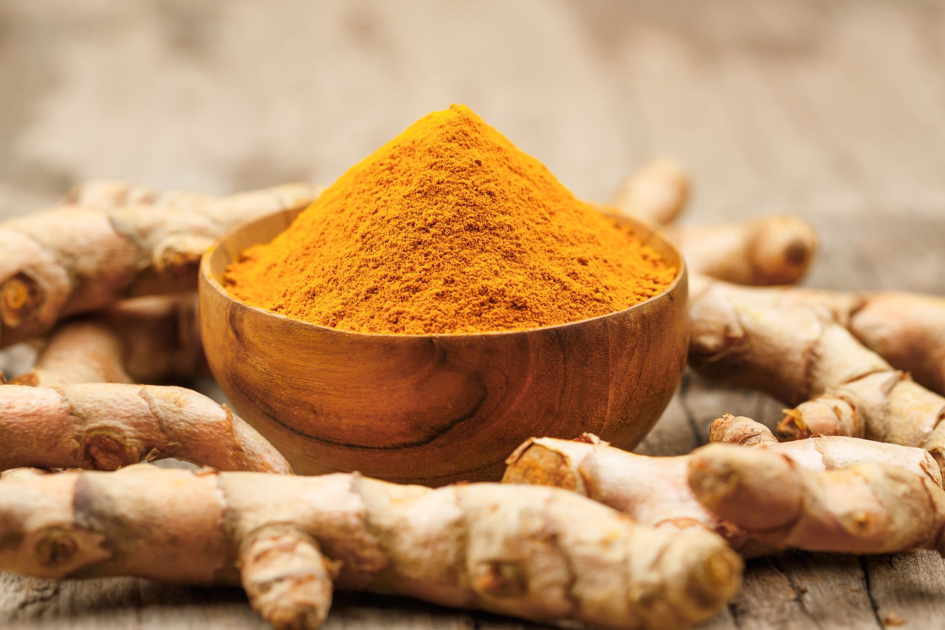 Wooden bowl with turmeric powder, surrounded by fresh turmeric roots, on a wooden surface.