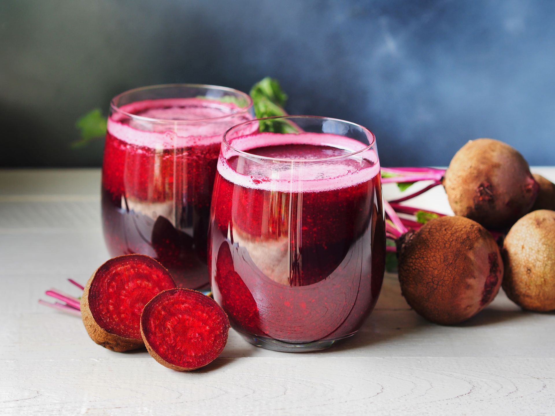 Two glasses of vibrant red beet juice, with beetroots and sliced beets on a light surface.