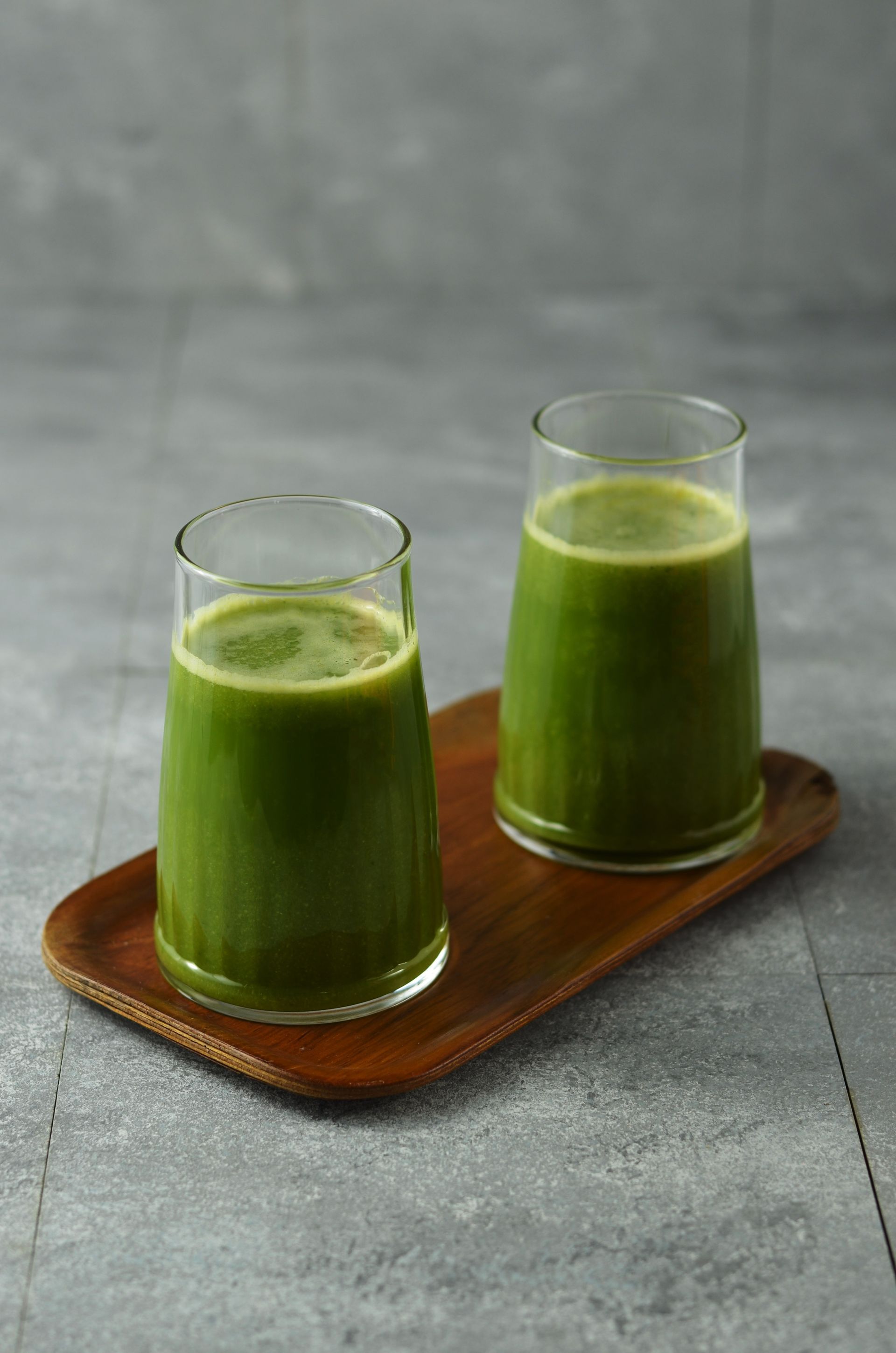 Two glasses of green juice on a wooden tray, against a gray background.