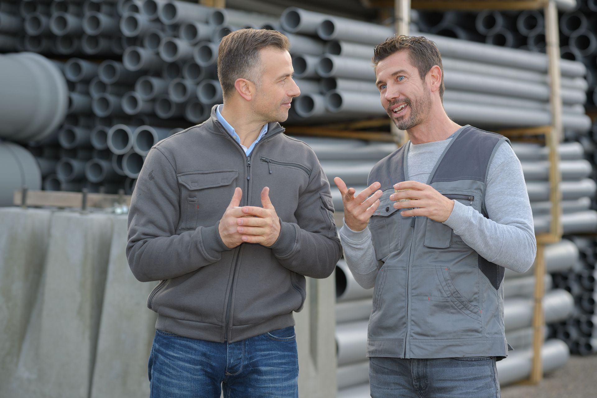 Two plumbing professionals discussing a project while standing near stacked pipes at a work site.
