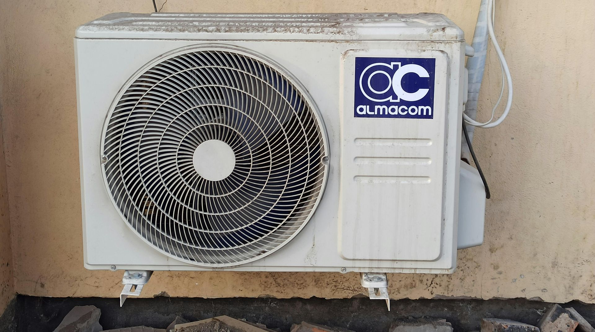 White outdoor air conditioner unit with a circular fan, mounted on a beige wall, with the brand name 
