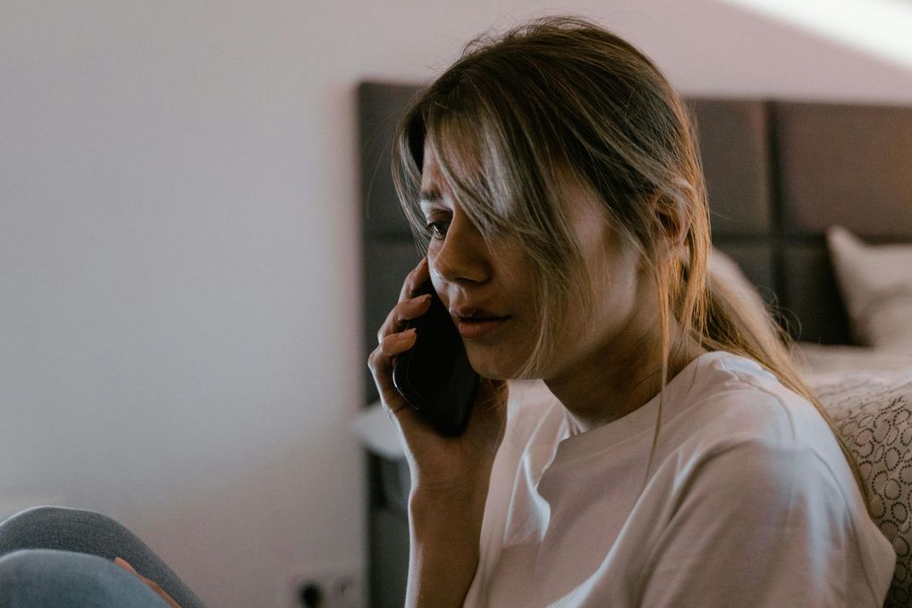 Woman on phone, leaning against a bed. She looks concerned, holding phone to ear. Room has natural light.