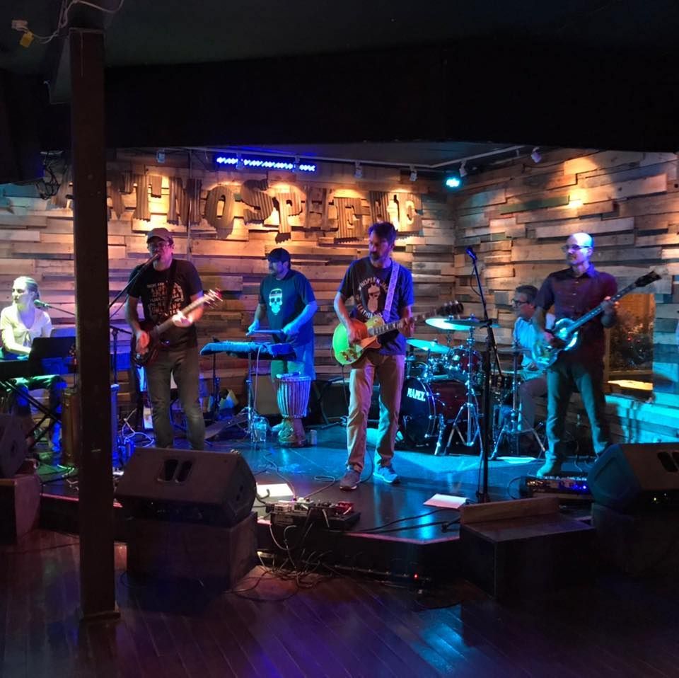 Band performing on stage at a venue with wood paneling. Musicians playing various instruments, colorful lighting.