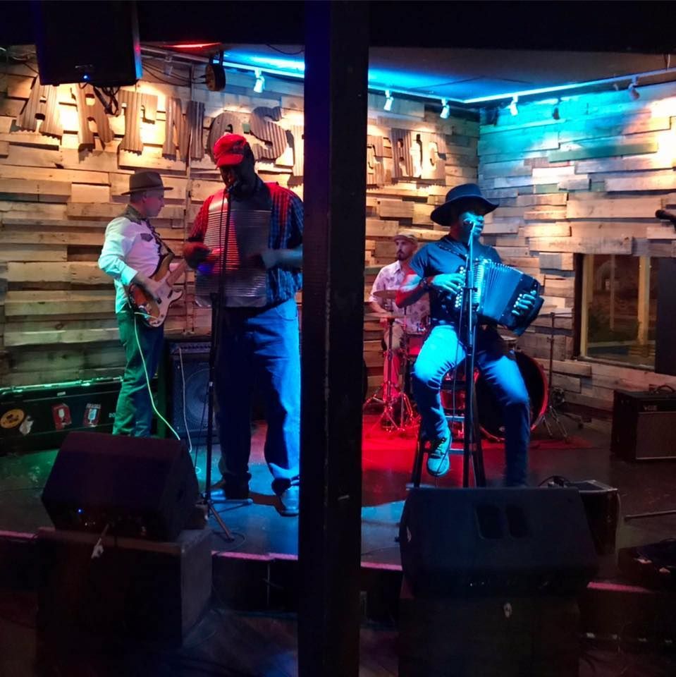 Band performing on stage: man with accordion, bass, drums, guitar. Stage with wood and blue/red lighting.