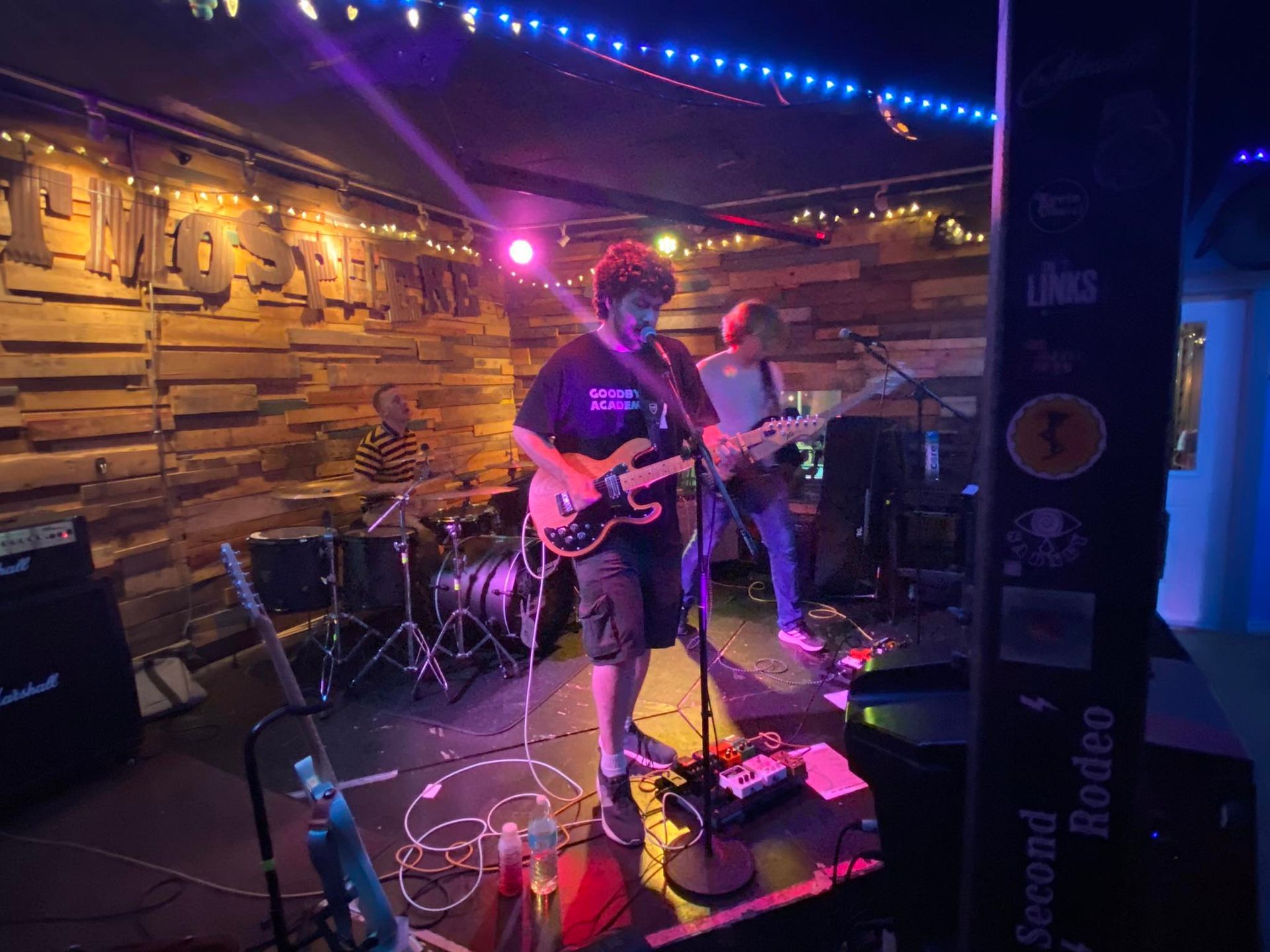 Band performing on stage at a bar, lit with blue and purple lights. Two guitarists, drums in background.
