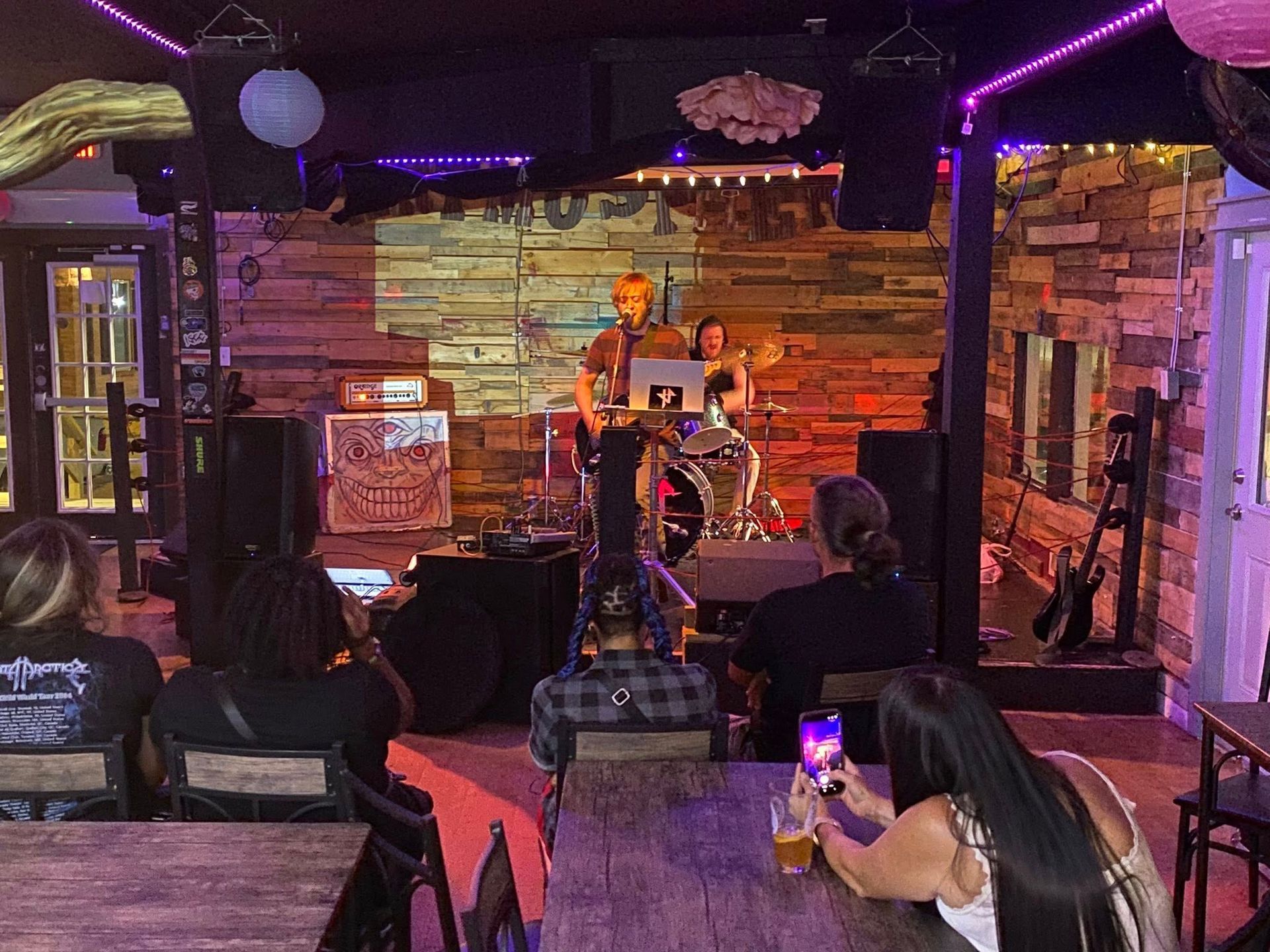 Band playing in a dimly lit bar. Audience watches. Stage has wood backdrop, purple and orange lights.