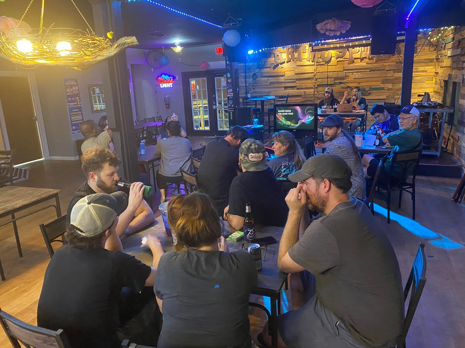 People gathered in a dimly lit bar, some seated at tables, others at the bar, enjoying drinks and conversation.