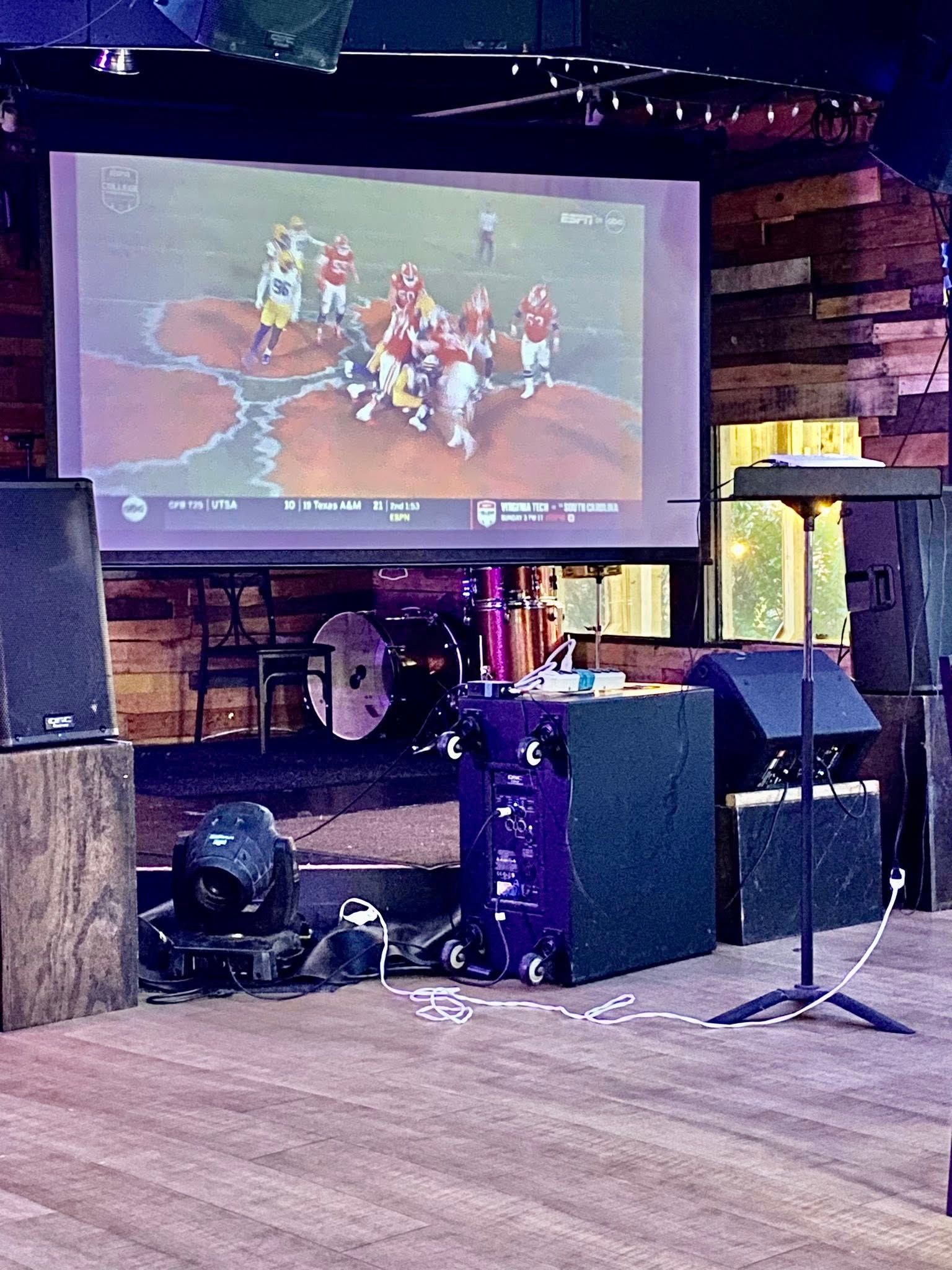 Large screen showing a football game in a dimly lit bar, surrounded by speakers and equipment.