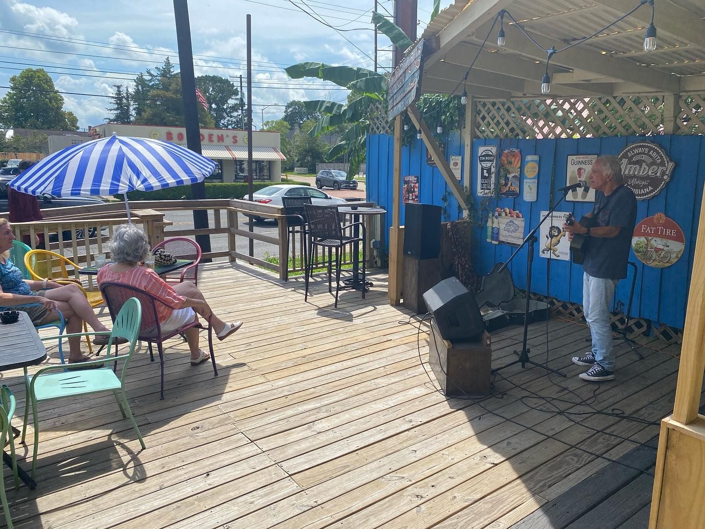 A musician performs outdoors on a wooden deck. People sit nearby. Blue sky.
