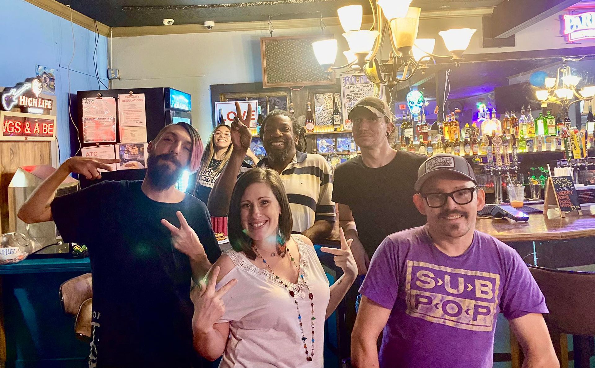 Group of people at a bar, smiling and giving peace signs. Brightly lit, colorful bottles behind the bar.