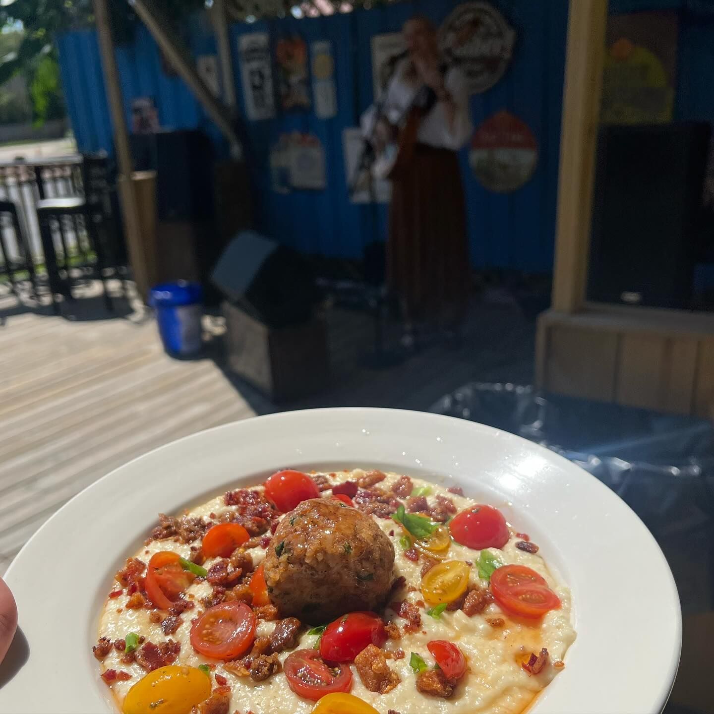 A plate of grits topped with tomatoes and a meatball, with a person singing on stage in the background.