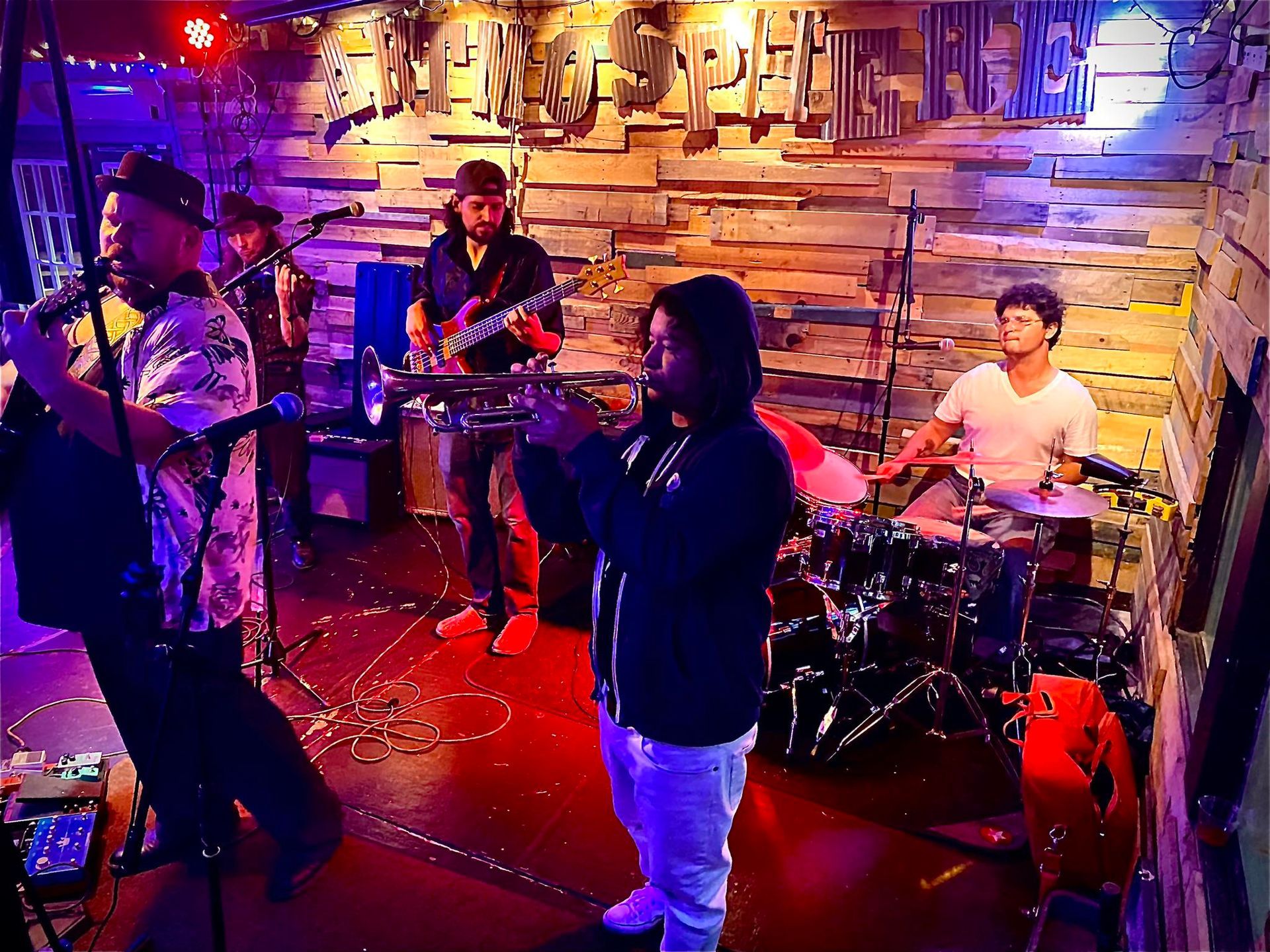 A band performs on stage. Musicians play saxophone, trumpet, guitar, drums, and bass in a dimly lit venue.