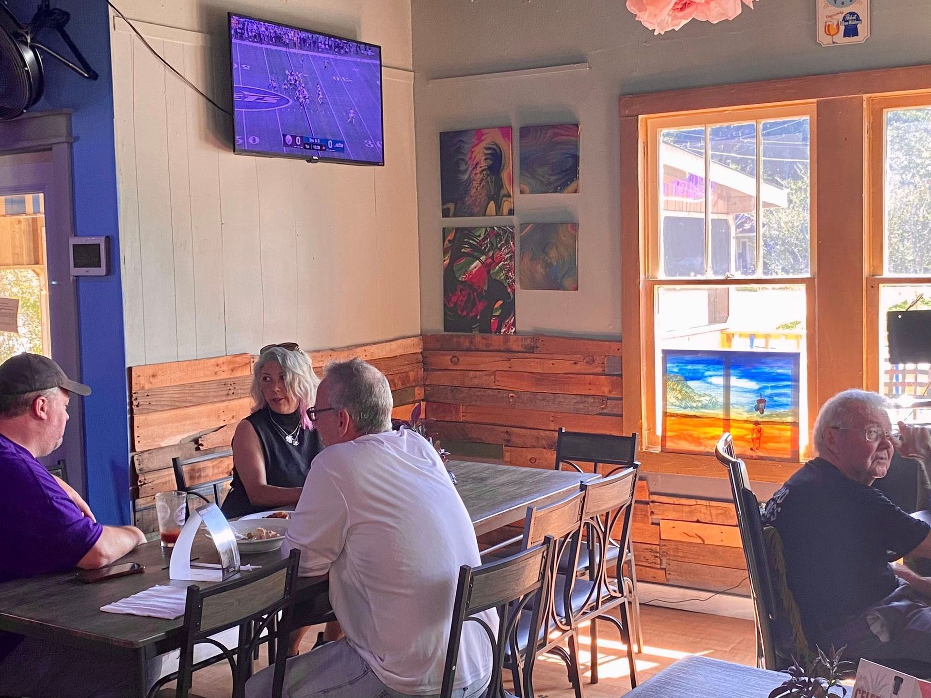 People seated around tables in a restaurant. A television and artwork are mounted on the walls. Sunlight streams in a window.