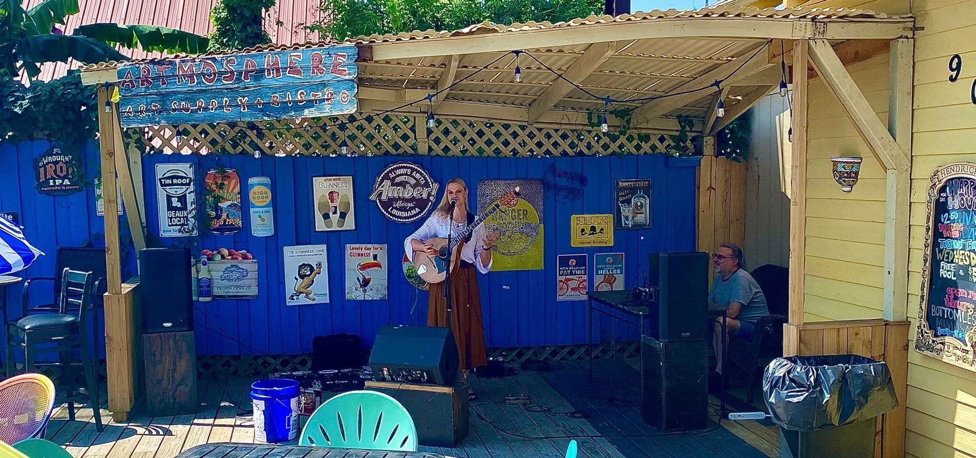 A person with a guitar performs on a small stage with a blue wall, posters, and audience member in the background.