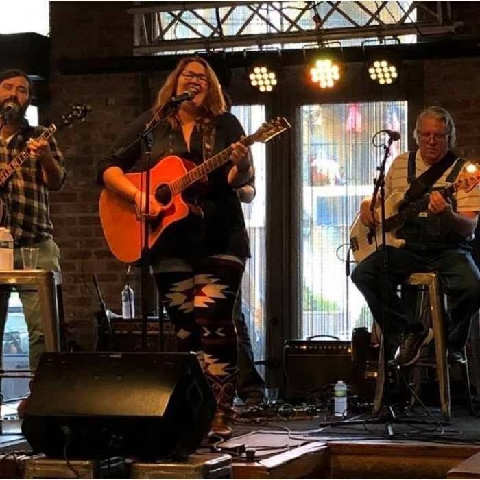 A band performs on stage: a woman singing and playing guitar, two men with instruments.
