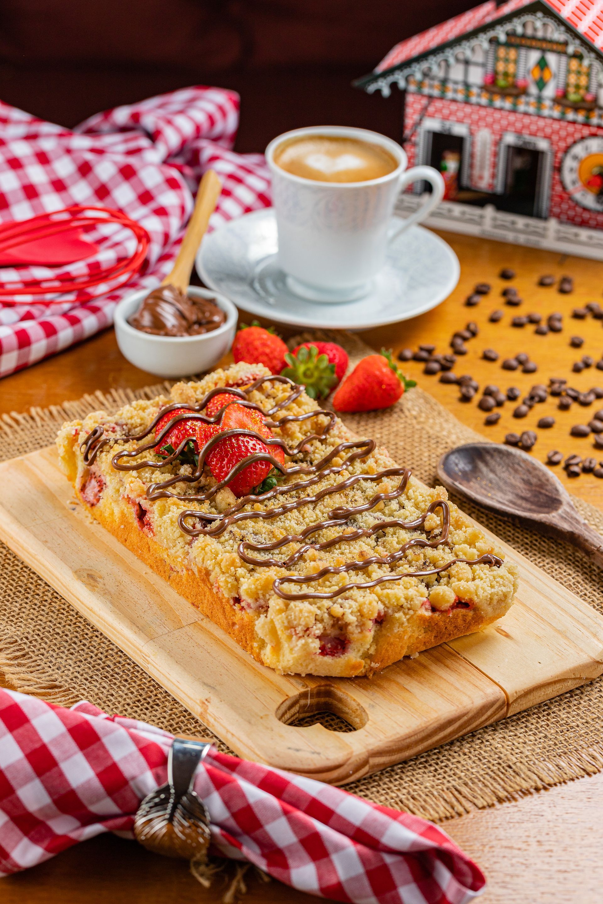 Um pedaço de cuca com morangos e calda de chocolate em uma tábua de madeira ao lado de uma xícara de café.