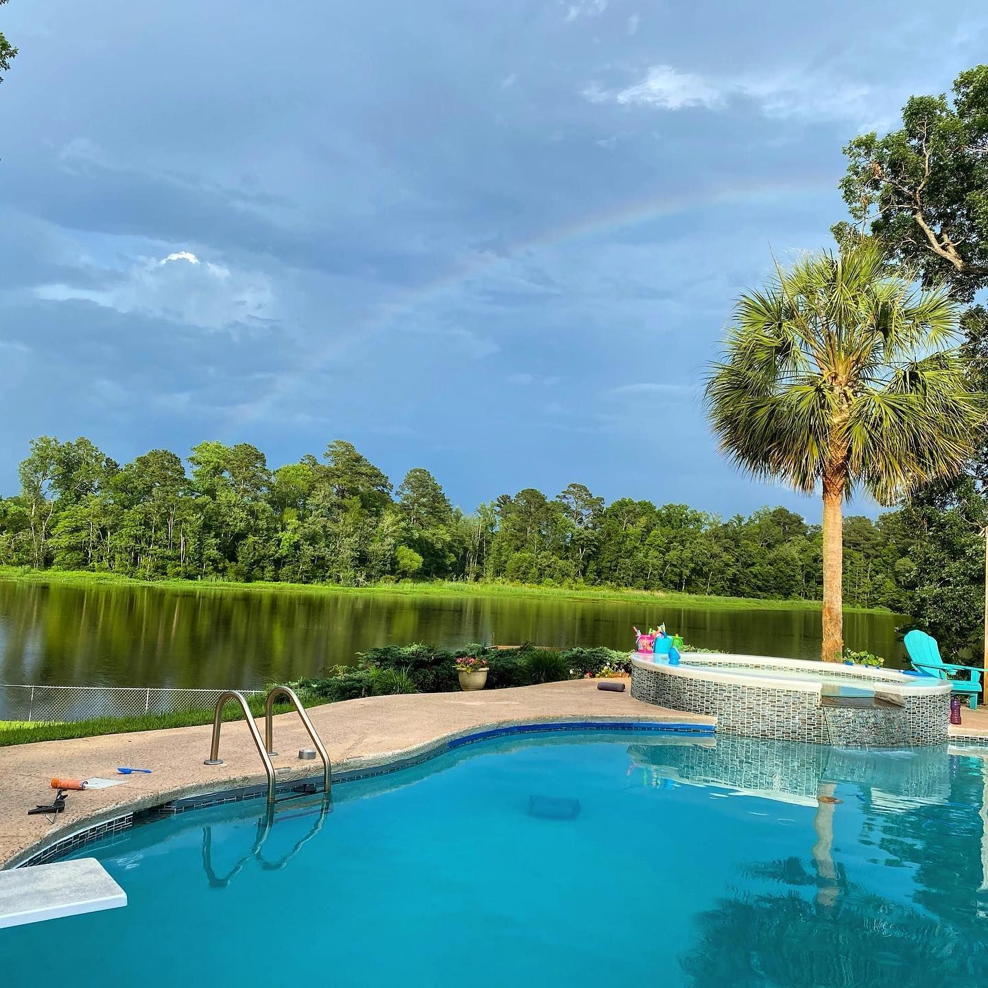 A swimming pool surrounded by trees and a lake with a rainbow in the sky