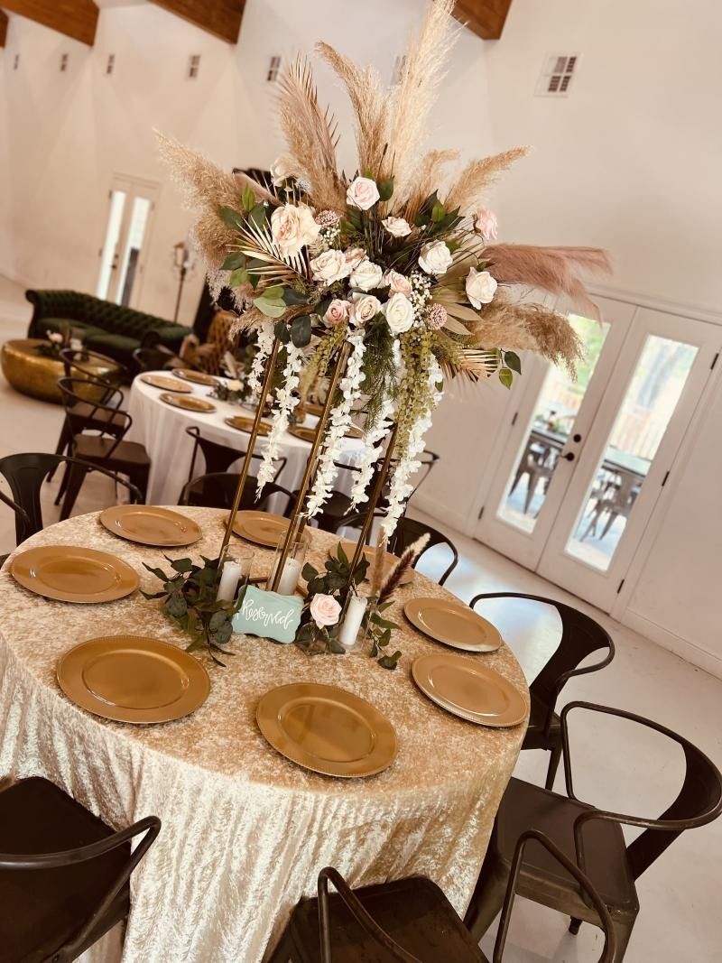 Elegant banquet table with gold plates, cream linens, and tall floral centerpiece in a bright reception hall