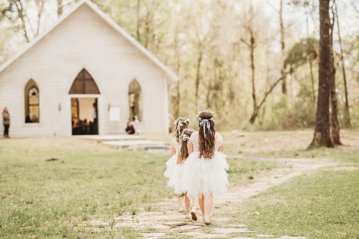 Two girls in white dresses walking toward a small chapel in a sunlit wooded clearing