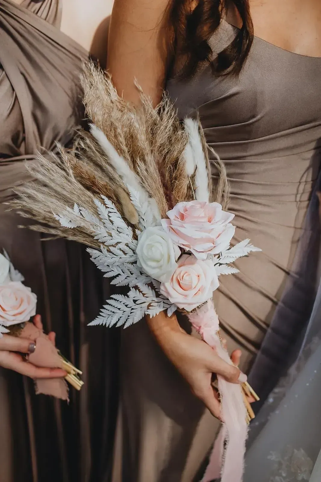 Three bridesmaids in taupe dresses holding pastel floral bouquets with pampas grass
