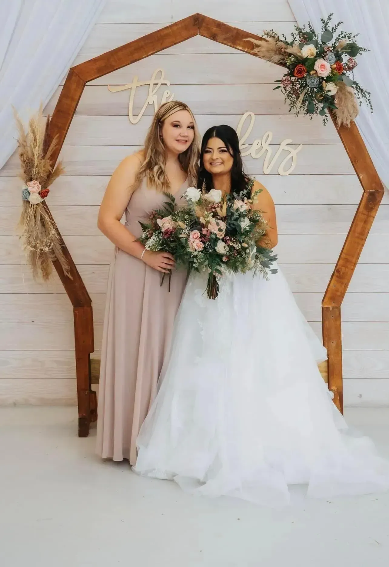 Two women in formal dresses posing with bouquets under a wooden hexagonal arch
