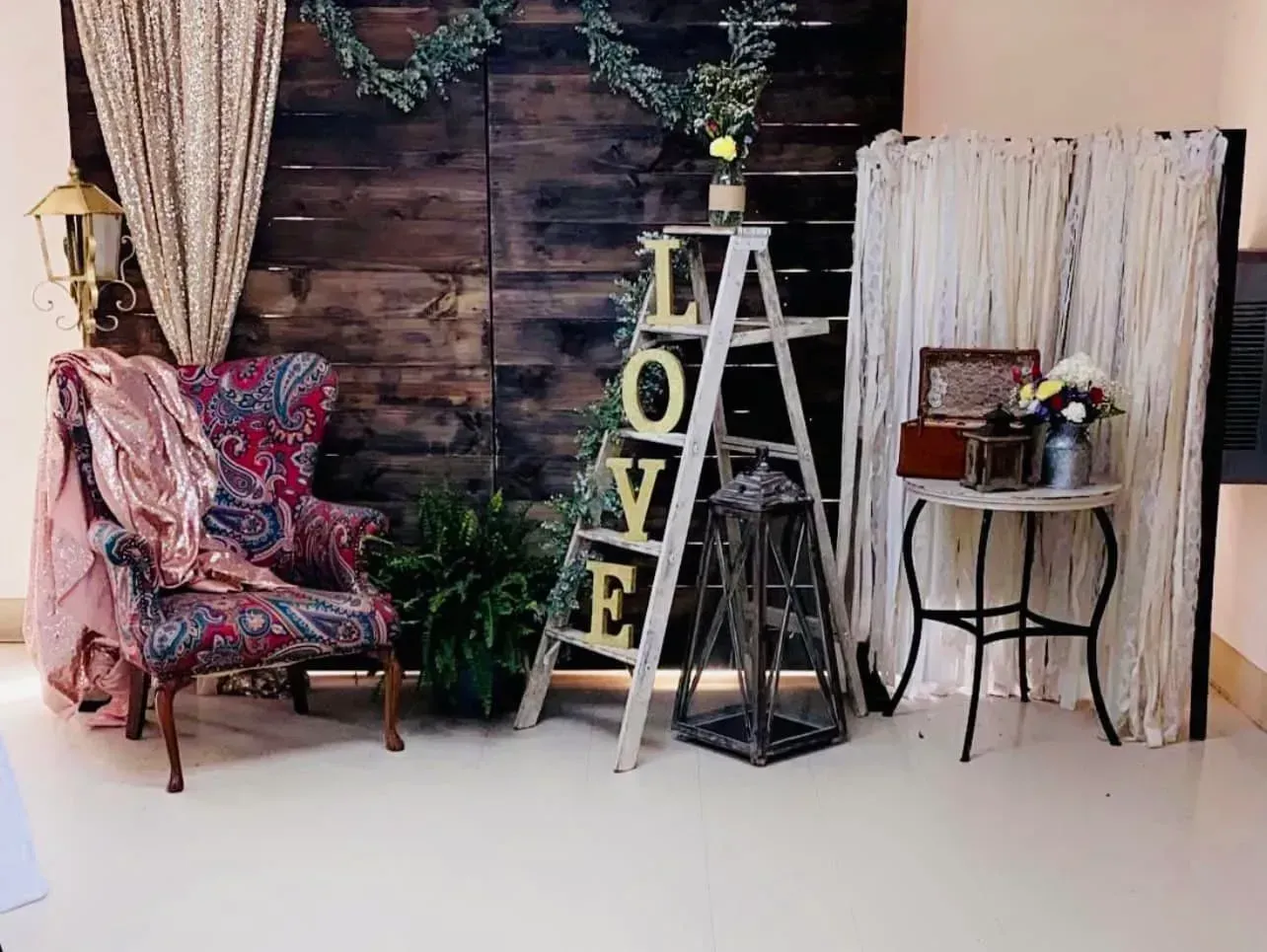 Cozy rustic room with floral armchair, ladder shelf, small table, and decorative greenery against wood backdrop
