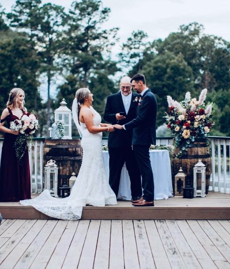 Outdoor wedding ceremony with bride and groom exchanging rings on a wooden deck, officiant and floral arrangements behind them