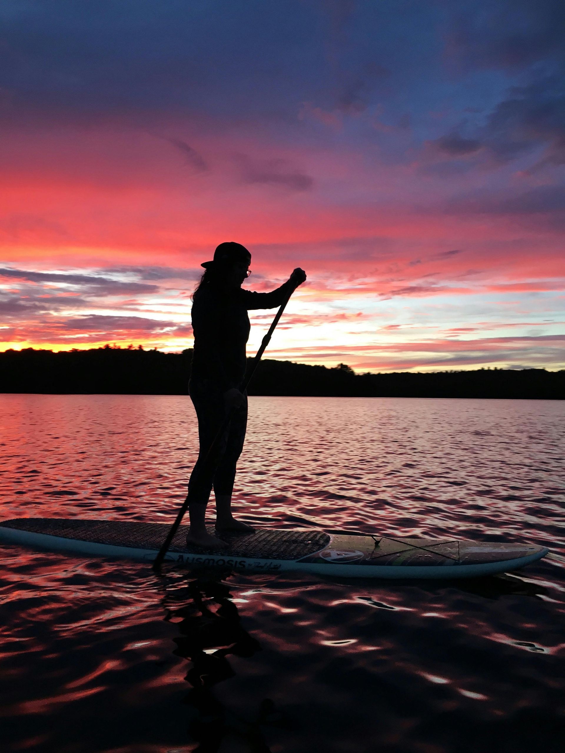 Person paddleboarding at sunset, silhouetted against vibrant red, orange and blue sky and water.