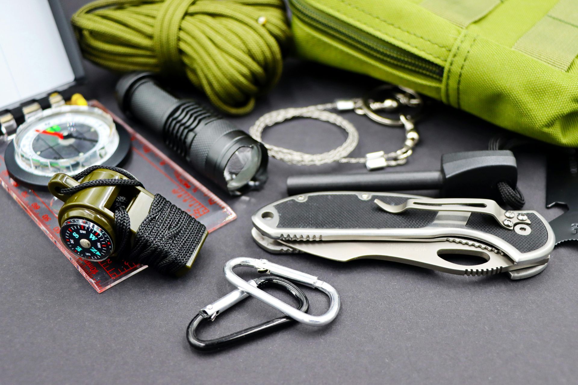 A survival kit laid out on a dark surface, featuring a folding knife, flashlight, compass, cord, and fire starter.