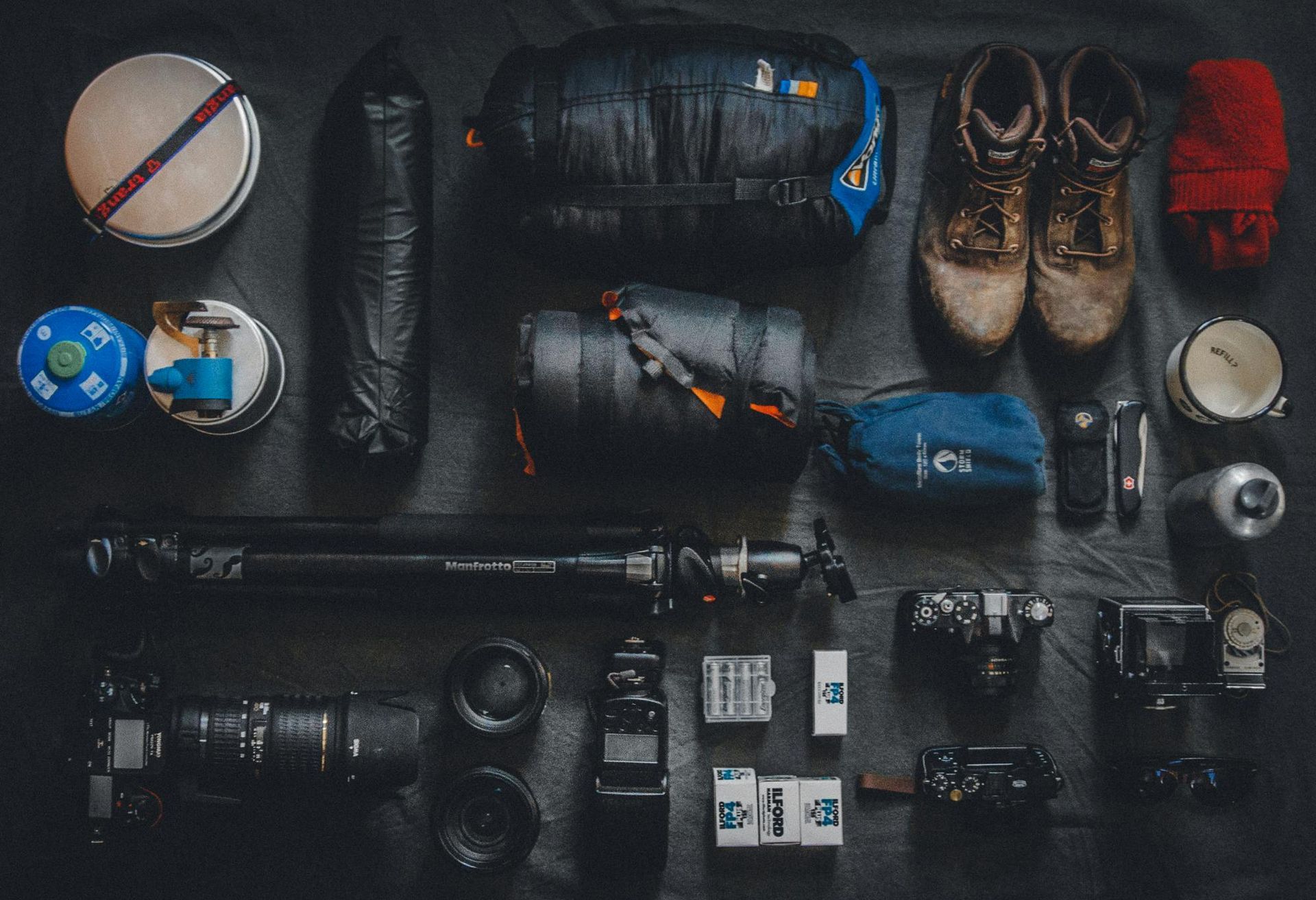 Top-down view of camping gear, boots, flashlight, camera, and tools arranged on a dark surface