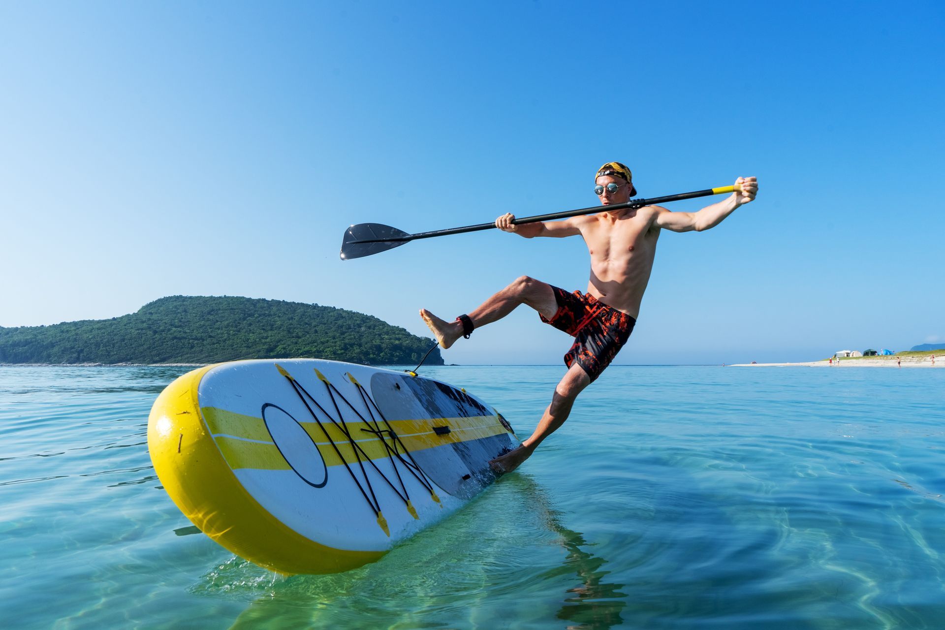 Man balancing on a paddleboard in the ocean, holding paddle, losing balance. Yellow and black paddleboard. Clear blue sky and water.