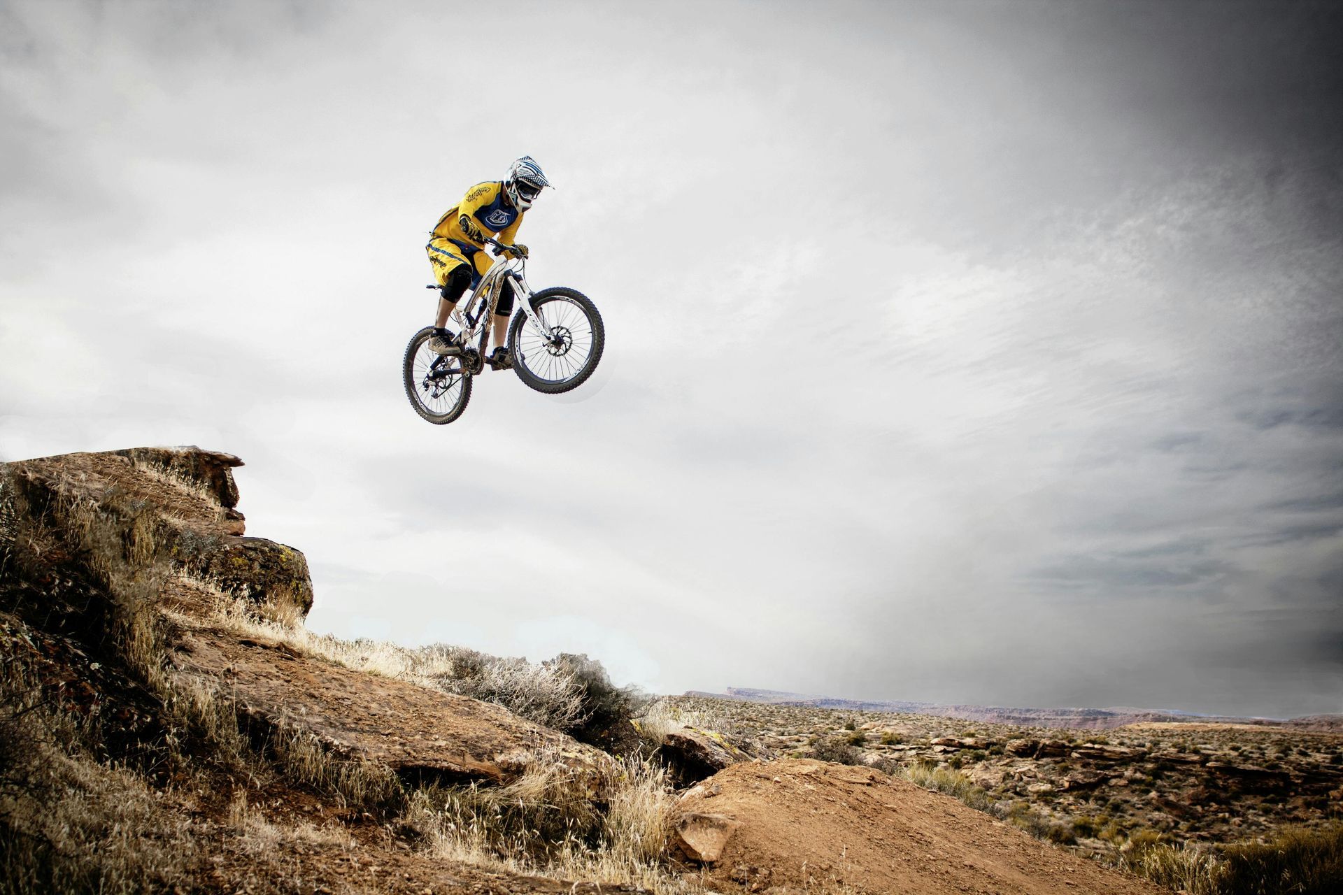 Mountain biker in mid-air over a dirt jump, wearing helmet and yellow gear against a cloudy sky.