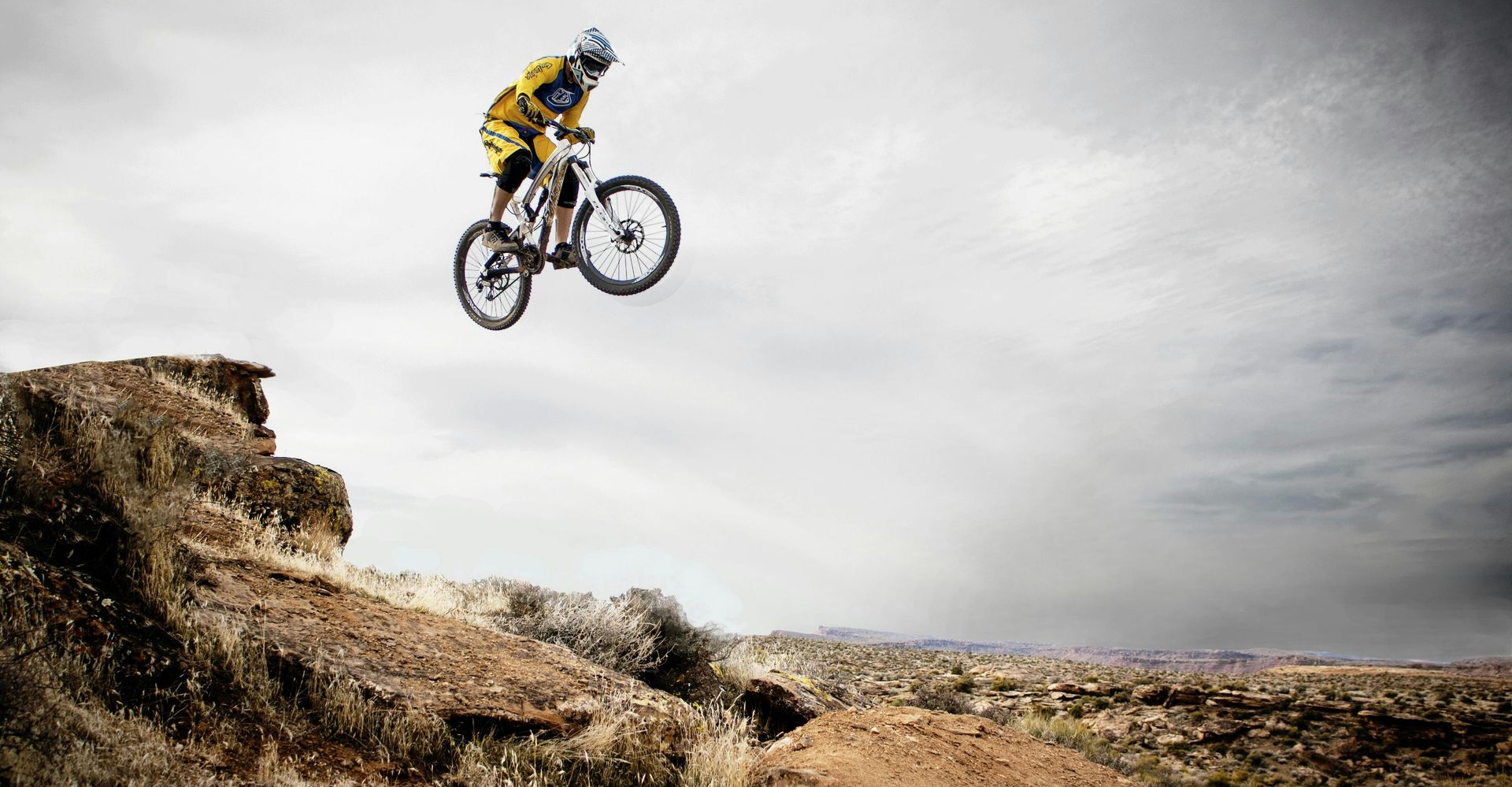 Mountain biker in mid-air, jumping off a cliff. Sky is cloudy. Brown terrain with sparse vegetation.