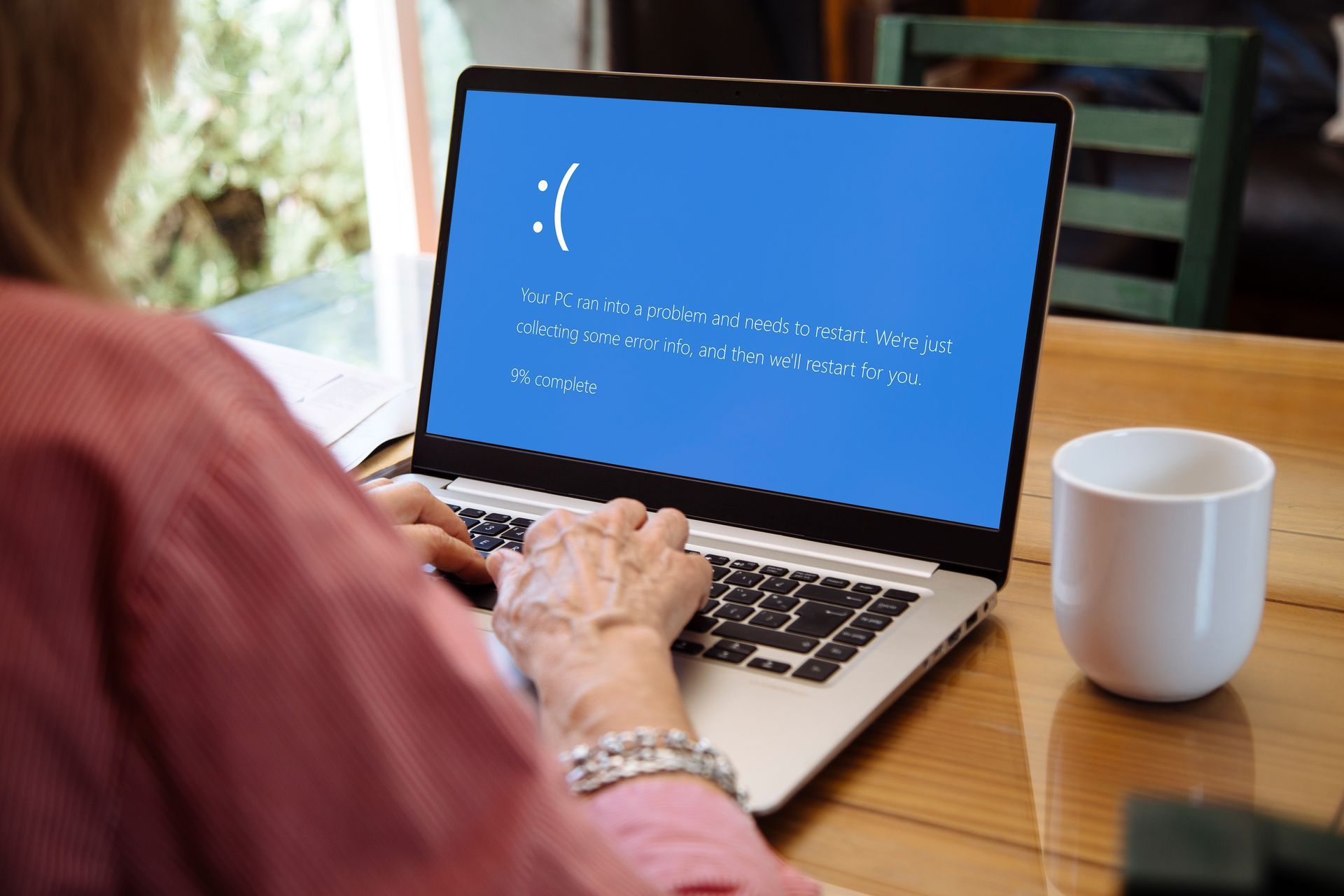 Woman using laptop with a blue screen of death, appearing distressed. Beside her is a white mug on a wooden table.