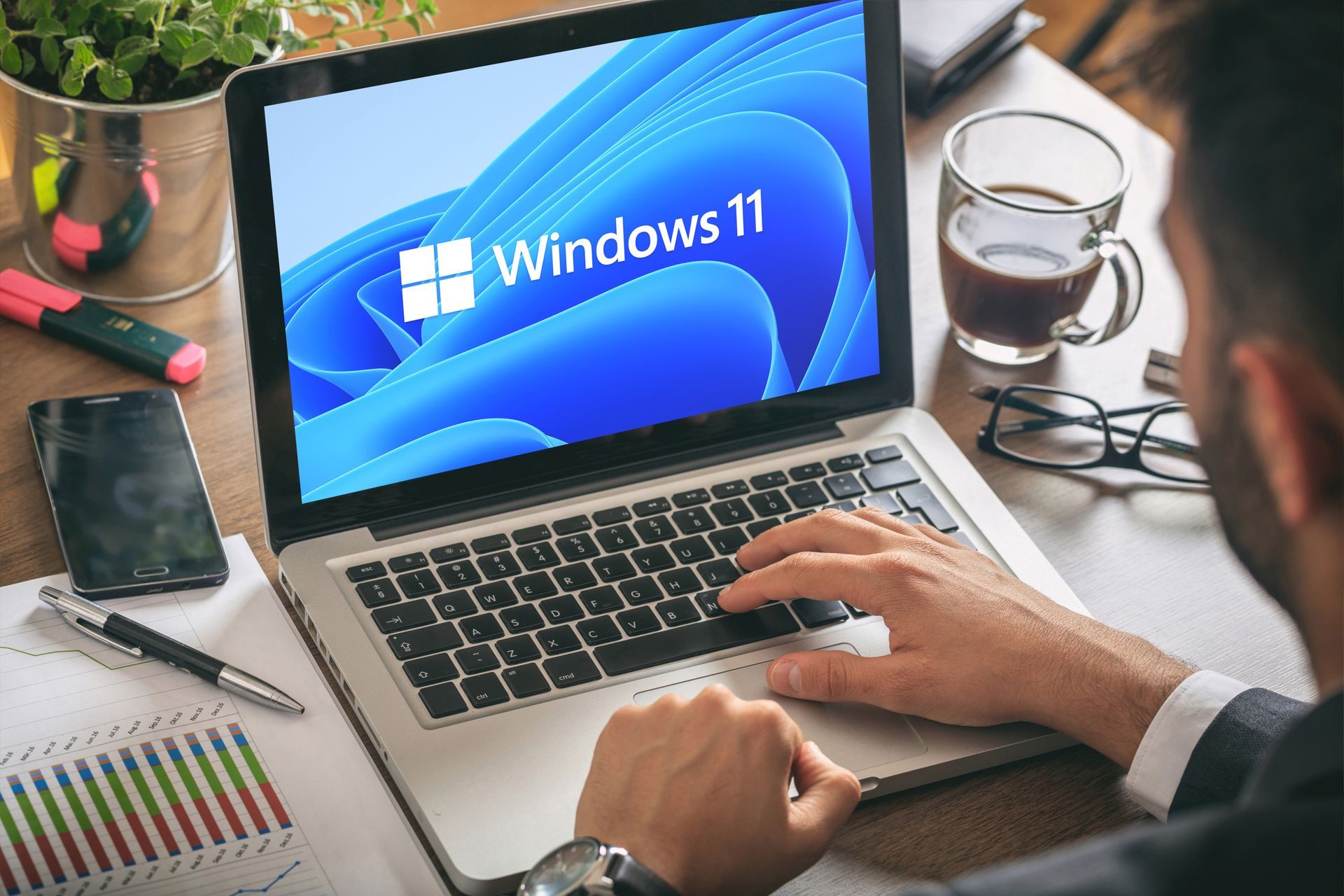 Person using a laptop displaying the Windows 11 logo on a desk with coffee, glasses, and a phone.