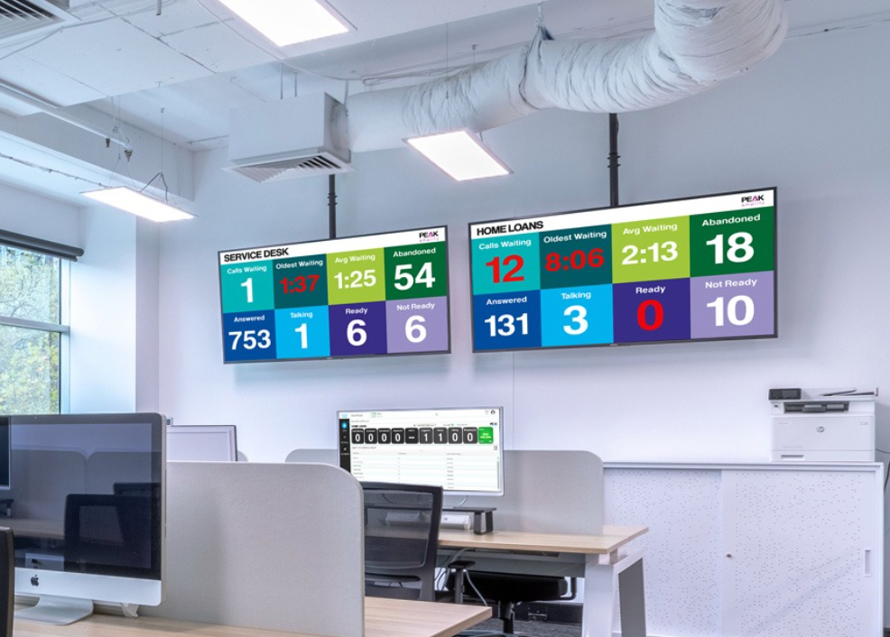Two large screens displaying data in a modern office above workstations, with one screen showing green, blue, red, and yellow data boxes.