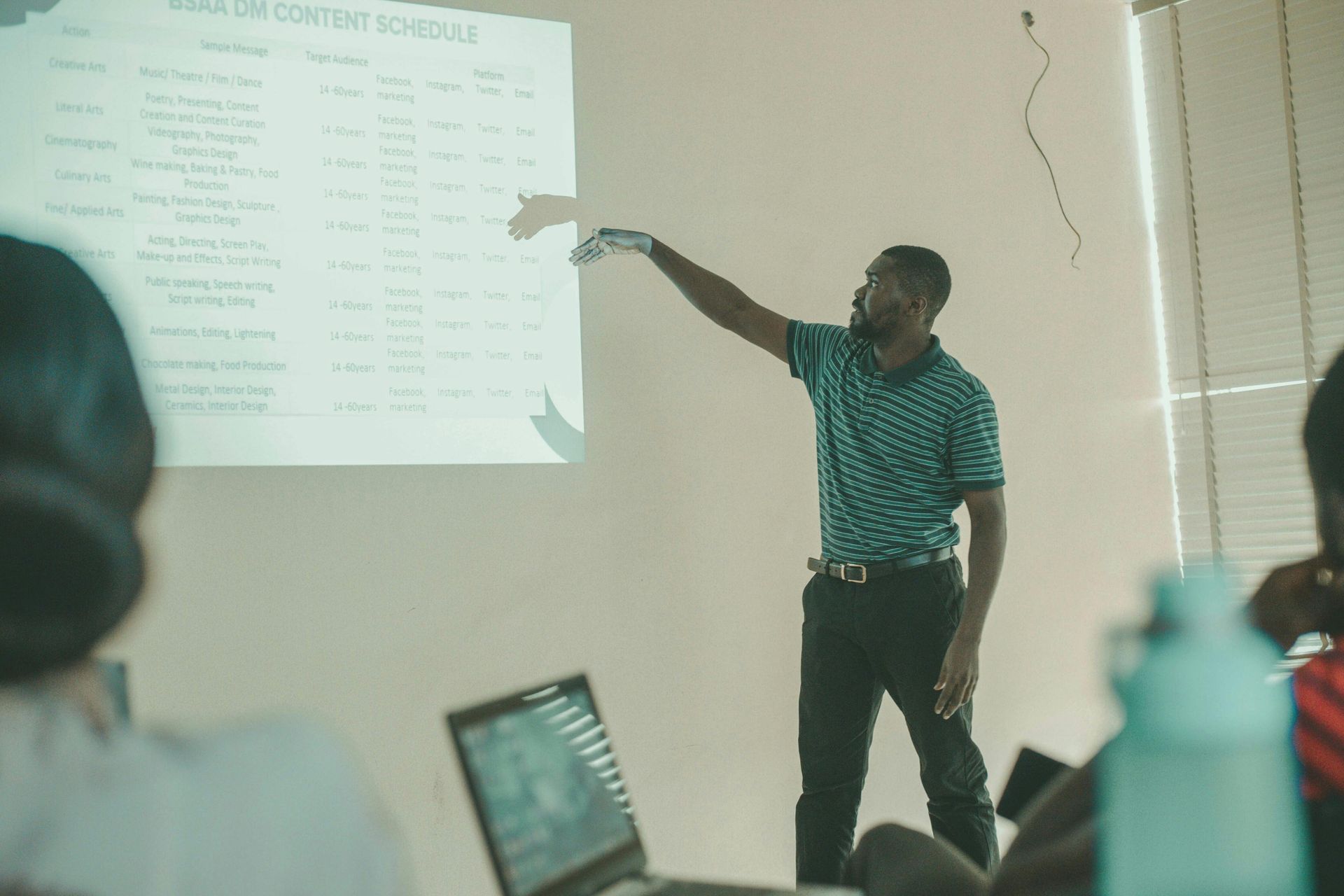 A man points at a projected schedule during a presentation. He wears a green shirt and dark pants.