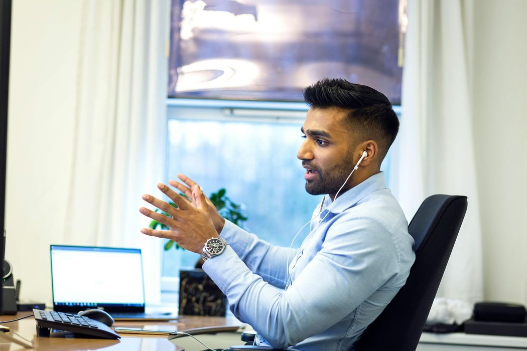 Man with earphones gesturing while speaking at desk with laptop and window.