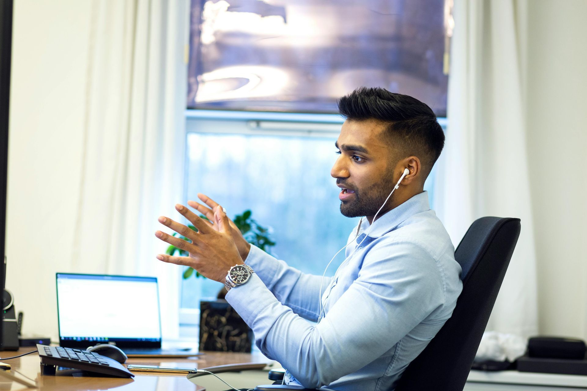 Man with earphones gesturing while speaking at desk with laptop and window.
