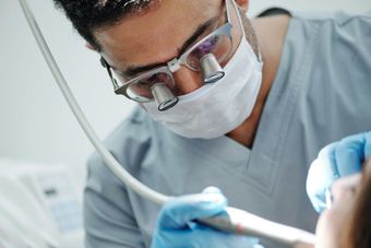 Dentist in mask and loupes, working on a patient's teeth. Blue gloves, setting is a dental office.